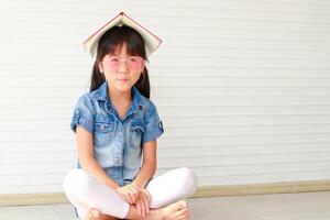 süß wenig asiatisch Mädchen haben Spaß, sie trägt Rosa Brille und setzt ein Buch auf ihr Kopf. sitzen auf das hölzern Fußboden im das Haus. Bildung Konzept Lernen zum Kinder. foto