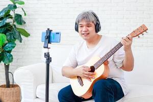 asiatisch Weiß behaart Mann spielen Musik- beim Zuhause genießen das Musik. lernen Musik- online durch Ihre Smartphone. Freizeit Aktivitäten. modern Kommunikation Neu Lebensstil foto