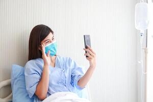 asiatisch weiblich geduldig tragen Maske im Krankenhaus geduldig Zimmer sie hält ein Smartphone online reden zu ihr Familie. Konzept von medizinisch Dienstleistungen. Patienten infiziert mit Coronavirus. Kopieren Raum foto