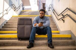 besorgt Mann mit ein Telefon und Koffer Sitzung auf ein Treppe beim das Eisenbahn Bahnhof. foto