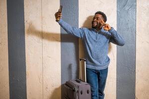glücklich Mann mit Koffer nehmen Selfie mit ein Telefon während gelehnt gegen ein Mauer im Passage von das Eisenbahn Bahnhof. Kopieren Raum auf Bild zum Ihre Inserat oder Text. foto