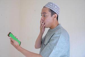 Moslem asiatisch Mann überrascht Geste Show das grüner Bildschirm auf Smartphone wann Ramadan Feier. das Foto ist geeignet zu verwenden zum Ramadhan Poster und Muslim Inhalt Medien.