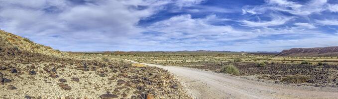 Panorama- Bild Über ein Kies Straße durch das Wüste mögen Steppe im Süd- Namibia unter ein Blau Himmel foto