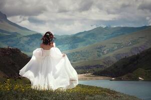 Braut im ein Weiß Hochzeit Kleid Spaziergänge im das Grün Berge auf das Gras, im das Hintergrund Dort sind Berge und ein See foto