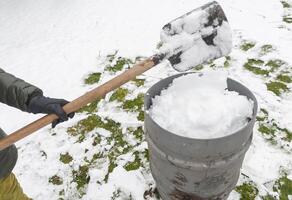 mittleren Alters Frau ist Sammeln Schnee im ein Fass mit ein Schaufel, natur Ressourcen foto
