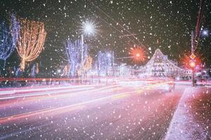 Nacht Winter Stadt mit Weihnachten oder Neu Jahr Dekorationen, Fichte und Spuren von Scheinwerfer von ziehen um Autos im ein Schneefall. Jahrgang Film ästhetisch. foto