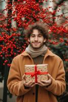 lächelnd jung Mann im ein Winter Mantel halten ein Weihnachten Geschenk mit ein rot Band draußen foto