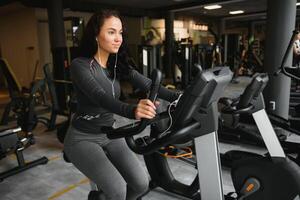 jung Frau mit Kopfhörer tun Übungen auf Schreibwaren Fahrrad im ein Fitnessstudio oder Fitness Center. jung sportlich Frau im Fitnessstudio Hör mal zu Musik- von Smartphone. Frauen tun Cardio Übungen foto