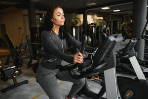 jung Frau mit Kopfhörer tun Übungen auf Schreibwaren Fahrrad im ein Fitnessstudio oder Fitness Center. jung sportlich Frau im Fitnessstudio Hör mal zu Musik- von Smartphone. Frauen tun Cardio Übungen foto