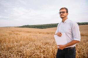 glücklich jung Farmer Ingenieur mit Notizbuch Stehen auf Weizen Feld während kombinieren Mähdrescher Arbeiten im Hintergrund. foto