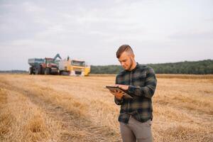 jung Agronom Mann Stehen auf Weizen Feld Überprüfung Qualität während kombinieren Mähdrescher Arbeiten foto