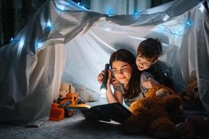 glücklich Familie Mutter und Kind Sohn lesen ein Buch mit ein Taschenlampe im ein Zelt beim heim. Familie Konzept. foto