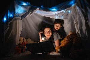 glücklich Familie Mutter und Kind Sohn lesen ein Buch mit ein Taschenlampe im ein Zelt beim heim. Familie Konzept. foto