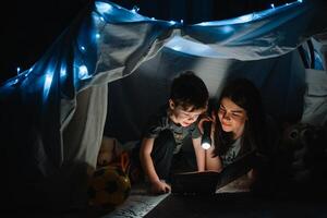 glücklich Familie Mutter und Kind Sohn lesen ein Buch mit ein Taschenlampe im ein Zelt beim heim. Familie Konzept. foto