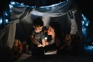 glücklich Familie Mutter und Kind Sohn lesen ein Buch mit ein Taschenlampe im ein Zelt beim heim. Familie Konzept. foto