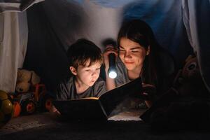 glücklich Familie Mutter und Kind Sohn lesen ein Buch mit ein Taschenlampe im ein Zelt beim heim. Familie Konzept. foto