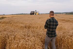 jung attraktiv Farmer mit Laptop Stehen im Weizen Feld mit kombinieren Mähdrescher im Hintergrund. foto