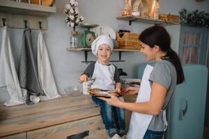 jung glücklich Mama und ihr Baby Koch Kekse beim Zuhause im das Küche. Weihnachten hausgemacht Lebkuchen. süß Junge mit Mutter im Weiß Uniform und Hut gekocht Schokolade Kekse foto