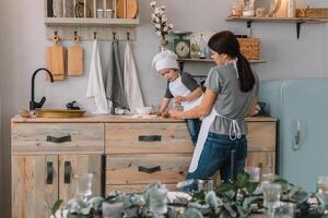 jung glücklich Mama und ihr Baby Koch Kekse beim Zuhause im das Küche. Weihnachten hausgemacht Lebkuchen. süß Junge mit Mutter im Weiß Uniform und Hut gekocht Schokolade Kekse foto