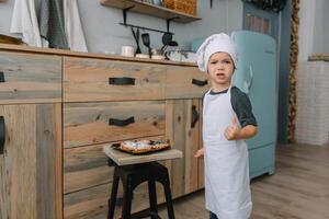 jung Junge süß auf das Küche Koch Koch im Weiß Uniform und Hut in der Nähe von Tisch. Weihnachten hausgemacht Lebkuchen. das Junge gekocht das Schokolade Kekse foto