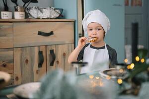 jung Junge süß auf das Küche Koch Koch im Weiß Uniform und Hut in der Nähe von Tisch. Weihnachten hausgemacht Lebkuchen. das Junge gekocht das Schokolade Kekse foto