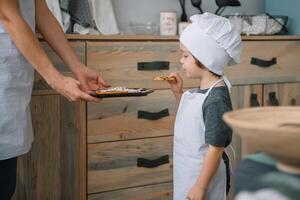 jung glücklich Mama und ihr Baby Koch Kekse beim Zuhause im das Küche. Weihnachten hausgemacht Lebkuchen. süß Junge mit Mutter im Weiß Uniform und Hut gekocht Schokolade Kekse foto