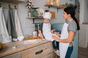 jung glücklich Mama und ihr Baby Koch Kekse beim Zuhause im das Küche. Weihnachten hausgemacht Lebkuchen. süß Junge mit Mutter im Weiß Uniform und Hut gekocht Schokolade Kekse. foto