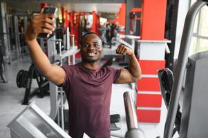 lächelnd jung Sportler zeigen Bizeps und nehmen Selfie mit Smartphone im Fitnessstudio. foto
