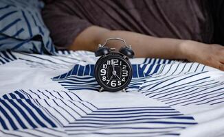 Frist und Zeit Verwaltung Konzept. schwarz Alarm Uhr auf Bett mit faul Menschen immer noch Schlafen im das Morgen foto
