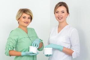 zwei Kosmetikerinnen mit Gläser von Wachs zum Enthaarung lächelnd gegen ein Weiß Hintergrund. natürlich Produkt zum Haar Entfernung. Kopieren Raum. foto