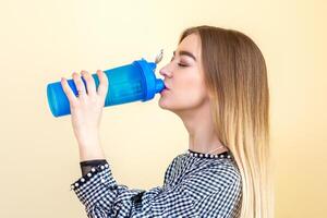 durstig Frau mit Blau Plastik Flasche gegen ein Licht Hintergrund. ein schön jung kaukasisch Geschäftsfrau im ein Bluse Trinken Wasser auf Gelb. foto