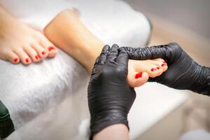 Fuß Massage mit feuchtigkeitsspendend und Peeling Sahne durch Fußpfleger Hände tragen schwarz Handschuhe, schließen hoch. foto
