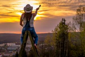 ein Frau im ein Arbeit Hose steht auf ein hölzern Leiter und Farben das Sonnenuntergang foto