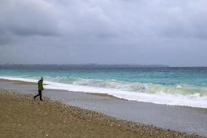 Antalya, Truthahn - - März 05, 2023. unkenntlich Mann ist Gehen in der Nähe von zu stürmisch Meer mit groß Wellen und Stadt auf Hintergrund. foto