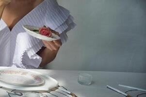 ein Frau, ein Mädchen im ein stilvoll Weiß Kleid hält ein Weiß Porzellan Untertasse mit ein Rosa Rose im ihr Hände. Tee Party, Morgen von das Braut. foto