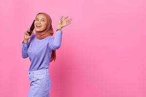 lächelnde junge asiatische frau, die mit handy spricht, während sie mit der handbewegung auf rosa hintergrund winkt