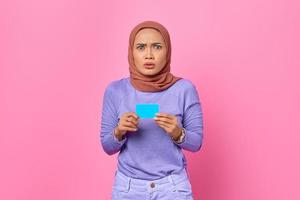Porträt einer schockierten jungen Asiatin, die Kreditkarte auf rosa Hintergrund zeigt