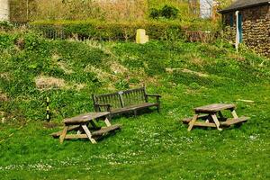 rustikal hölzern Bänke und ein Tabelle auf ein üppig Grün grasig Hang, mit ein Stein Gebäude und Vegetation im das Hintergrund, abbilden ein heiter draussen Rahmen im York, Norden Yorkshire, England. foto