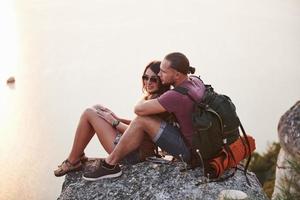 umarmendes Paar mit Rucksack, das oben auf dem Felsenberg sitzt und die Aussicht auf einen Fluss oder See genießt. Reisen entlang von Bergen und Küste, Freiheit und aktives Lifestyle-Konzept foto