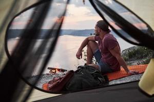 Blick vom Zelt des Reisenden mit Rucksack, der auf dem Berg sitzt und die Aussicht auf die Küste eines Flusses oder Sees genießt. Reisen entlang von Bergen und Küste, Freiheit und aktives Lifestyle-Konzept foto