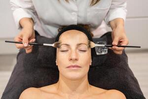 Ernte Kosmetikerin mit Bürsten zum Gesicht von weiblich Klient im Schönheit Salon foto