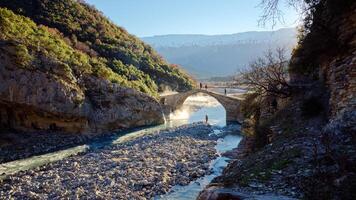 Menschen Kreuzung das kadiut Brücke im Benje, Erlaubnis, Albanien. uralt Stein Brücke. schön natürlich Wunder. Ferien und Freizeit Zeit. Fischer Angeln im das Fluss. natürlich Thermal- Bad. foto