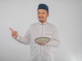 Moslem asiatisch Mann lächelnd und zeigen zu das richtig Seite während halten leeren Abendessen Teller foto