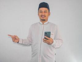 Moslem asiatisch Mann lächelnd glücklich während halten Handy, Mobiltelefon Telefon und zeigen zu das richtig Seite foto