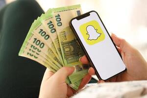 Kiew, Ukraine - - April 1, 2024 Snapchat Symbol auf Smartphone Bildschirm und Geld im weiblich Hand foto