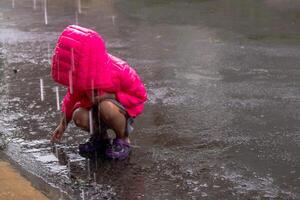 ein wenig asiatisch Mädchen im ein duckte sich Position tragen ein Rosa Jacke und spielen im das Regen foto