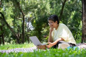 glücklich asiatisch Senior Frau mit Laptop zum Arbeiten online draußen Büro. Alten Pensionierung mit Laptop zum Lernen Neu Fertigkeit und freiberuflich Arbeiten im Park, Senior Lebensstil mit Technologie foto