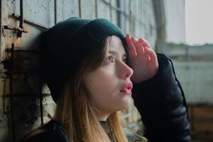 Porträt von ein schön Teenager Mädchen im ein schwarz Winter Jacke und Hut gegen das Hintergrund von ein unvollendet, zerstört und verlassen Gebäude Mauer. städtisch Konzept. Nahansicht. foto