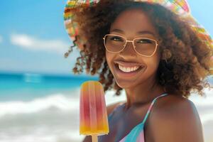 Porträt von ein jung lächelnd afrikanisch amerikanisch Frau Essen ein Eis am Stiel Eis Sahne auf heiß Sommer- Tag beim das Strand foto