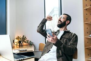 Ja, es funktioniert. heiter bärtig Mann im Brille freut sich im ein Cafe mit ein Handy, Mobiltelefon Telefon, Herstellung ein Gewinner Geste. entfernt arbeiten. Kaffee Geschäft, Fernbedienung arbeiten, freiberuflich foto